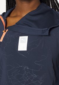 Persona con capelli intrecciati che indossa una giacca sportiva blu navy con un leggero motivo a mappa e un piccolo logo Puma vicino al colletto.