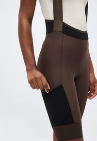 Άτομο που φορά καφέ και μαύρο ποδηλατικό σορτς με τιράντες, τραβώντας το ύφασμα της πλευρικής τσέπης.