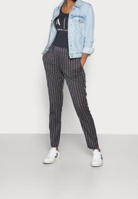 Chaqueta de mezclilla clara sobre una camiseta sin mangas azul marino. Pantalones rayados en negro y color óxido, combinados con zapatillas blancas con detalles en negro.