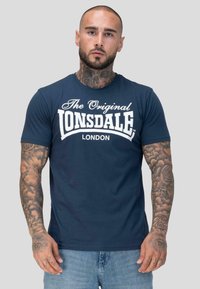 Mees, kellel on lühike habe ja tätoveeringud kaelal ning käsivartel, kannab tumesinist T-särki "Lonsdale London" ning helesiniseid teksaseid, seistes ühevärvilise tausta ees.