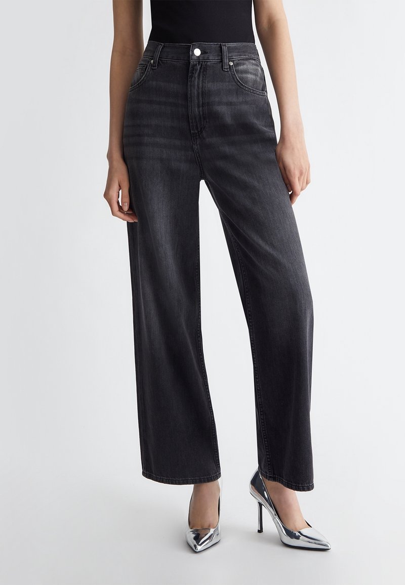 Femme portant un jean noir taille haute à jambes larges et des talons hauts argentés pointus, debout devant un fond clair uni.