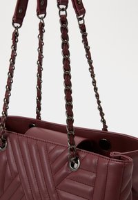 Sac à main bordeaux avec texture matelassée, doté de deux anses : une lisse et une en chaîne, œillets en métal, et un intérieur spacieux.