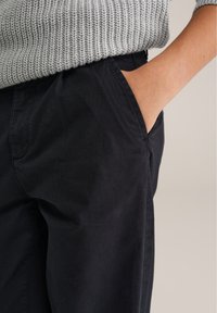 Osoba v čiernych nohaviciach s pravou rukou ležérne vloženou v prednej vrecku, v kombinácii so svetlomodrým pleteným sveterom.