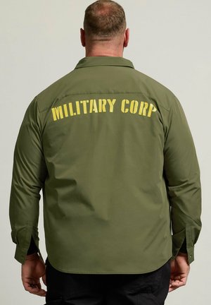 Mand med kort hår iført en olivengrøn jakke med gul "MILITARY CORP" tekst på ryggen, stående mod en ensfarvet baggrund.