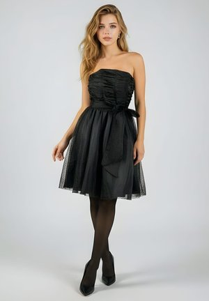 Joven con cabello largo y rubio, vestida con un vestido negro sin tirantes hasta la rodilla, medias negras y tacones negros, de pie frente a un fondo liso.