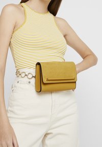 Borsa a pochette gialla in suede con una patta, bordi neri e una striscia orizzontale decorativa. Indossata su uno sfondo di un abito bianco.