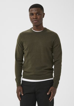 Mees kannab oliivirohelist pika varruka jahekotti valge särgi kohal, seisab käed kõhul rahulikult all tavalise tausta ees.