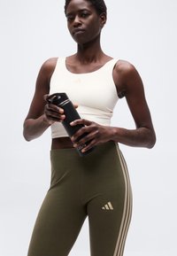 Αθλητικό άτομο που φοράει λευκό αθλητικό τοπ και χακί κολάν κρατάει μαύρο παγούρι Casall σε απλό ανοιχτόχρωμο φόντο.