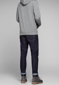 Sudadera con capucha gris, jeans azul oscuro con puños enrollados y botas grises. El atuendo casual presenta un diseño minimalista y un corte relajado.