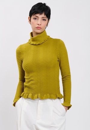 Frau mit kurzen dunklen Haaren, die einen strukturierten senfgelben Rollkragenpullover und weiße Hose trägt, die Hände in den Taschen vor einem schlichten Hintergrund.