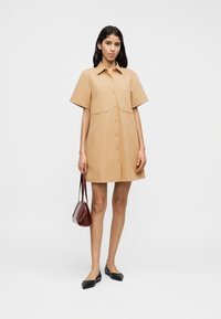 Μπεζ φόρεμα-πουκάμισο με κοντά μανίκια, κουμπιά μπροστά και δύο τσέπες στο στήθος. Συνδυασμένο με μαύρα φλατ παπούτσια και μικρή μπορντό τσάντα.