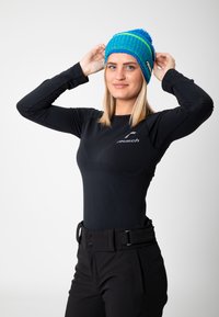 Knit beanie in blue and green with a pom-pom on top; fitted black long-sleeve top; black pants with a flat waistband and button details.