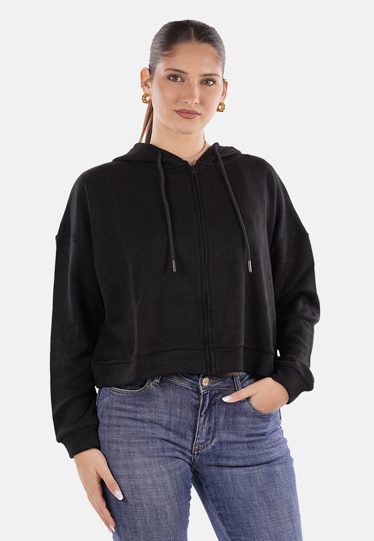 Giovane donna con capelli raccolti che indossa orecchini a cerchio d'oro, una felpa nera con zip e jeans blu, con una mano in tasca.