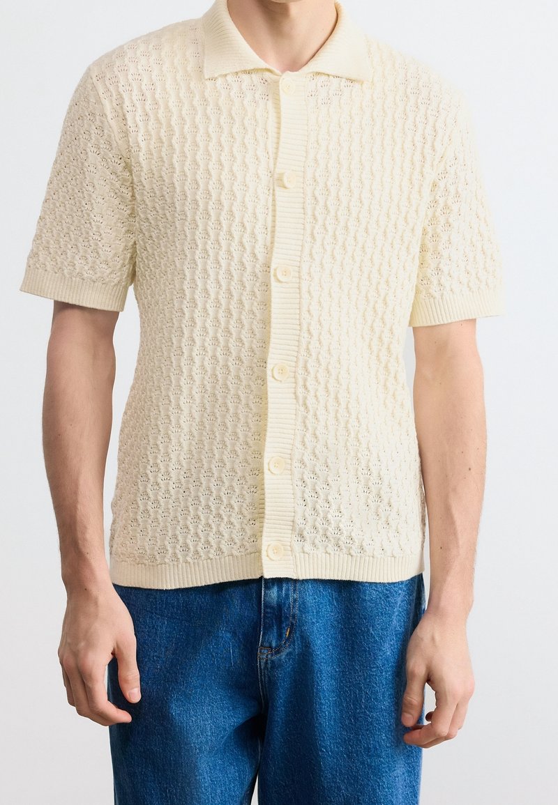 Homme portant un cardigan en maille crème à manches courtes avec des boutons et un col, associé à un jean bleu en denim.