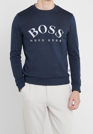 Mężczyzna w granatowej bluzie Hugo Boss z białym logo i jasnobeżowych spodniach, stojący z jedną ręką w kieszeni.