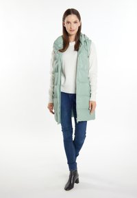 Mintgrön pufferväst med huva, dragkedja fram och tryckknappar, paired med en ljus tröja och mörka slim fit-jeans. Svarta klackboots.