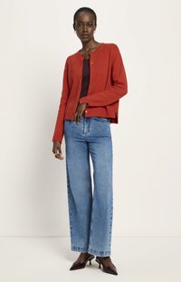Frau in rotem, geknöpftem Cardigan, burgunderfarbenem Oberteil, weitgeschnittenen blauen Jeans und burgunderfarbenen spitzen Pumps, die vor einem einfarbigen Hintergrund steht.