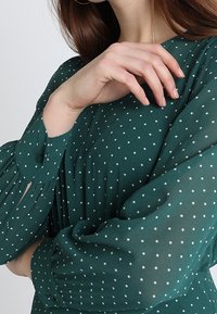 Blouse verte à pois avec des manches bouffantes, en tissu léger. Elle présente un détail de boutons au niveau des poignets et une coupe ample et fluide.