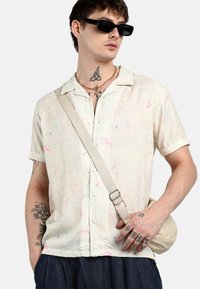 Camicia a maniche corte in tessuto beige chiaro con motivi di note musicali colorate; abbinata a una borsa a tracolla neutra e pantaloni scuri.