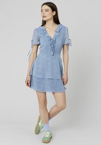 Robe courte bleue à motif floral avec des volants, des manches courtes et une jupe à niveaux. Associée à des baskets vertes et des chaussettes bleu clair.