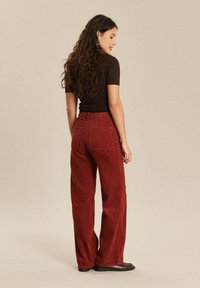 Pantalons larges en velours côtelé rouge avec des nervures texturées, taille haute et deux poches arrière. Le tissu présente un motif à côtes.