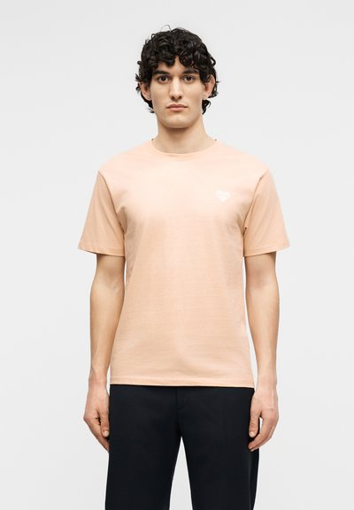 Νεαρός άνδρας με σγουρά σκούρα μαλλιά, που φορεί κοντομάνικη φανέλα χρώματος ανοιχτό ροδακινί και μαύρο παντελόνι, στέκεται μπροστά σε απλό φωτεινό φόντο.