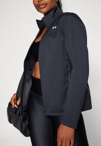 Črna atletska jakna z visokim ovratnikom, zadrgo spredaj in logotipom Under Armour. Ima eleganten, gladek teksturo in prilegajočo obliko.