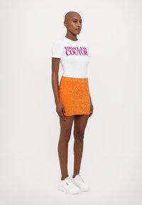 Fehér, testhezálló póló a "Versace Jeans Couture" logójával, párosítva egy rövid, élénk narancssárga gyöngyös szoknyával és fehér, vastag talpú sportcipőkkel.