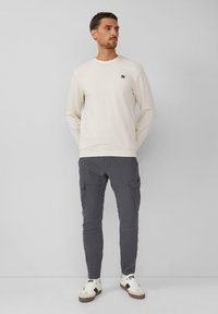 Lichtbeige sweatshirt met een klein logo, gecombineerd met donkergrijze cargobroek met zijzakken, en witte sneakers met bruine accenten.
