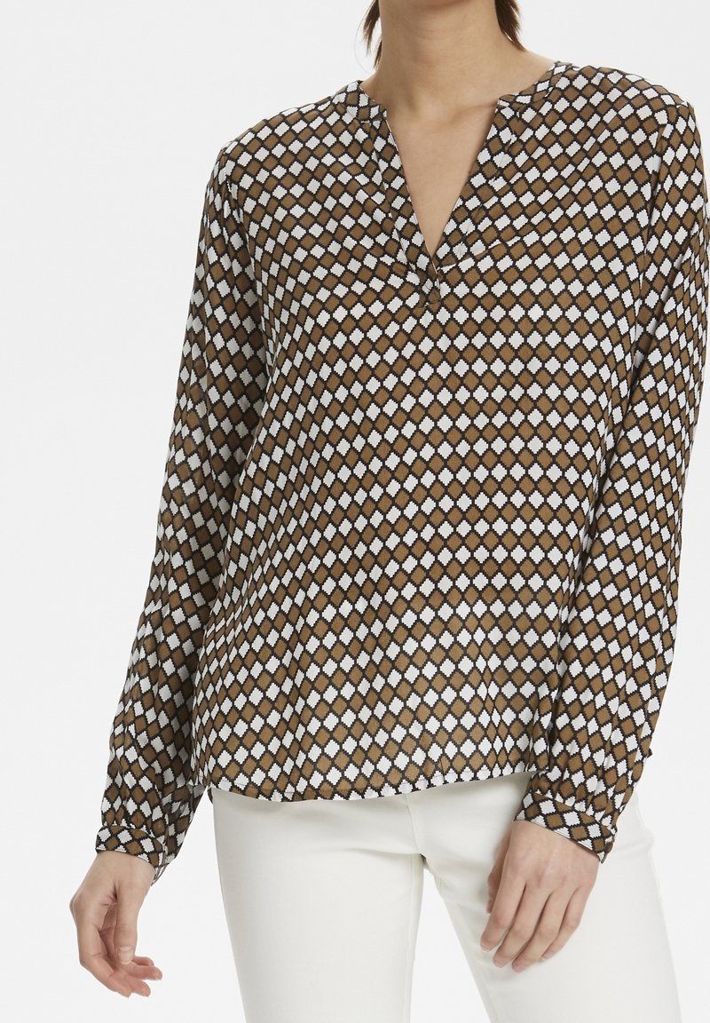 Femme portant un chemisier à manches longues avec un motif géométrique marron et blanc, associé à un pantalon blanc, debout devant un fond uni.