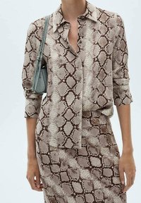 Blusa a maniche lunghe marrone e crema con colletto e chiusura con bottoni, caratterizzata da un motivo in pelle di serpente. Il modello porta una piccola borsa grigia.