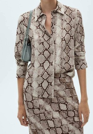 Blusa a maniche lunghe marrone e crema con colletto e chiusura con bottoni, caratterizzata da un motivo in pelle di serpente. Il modello porta una piccola borsa grigia.