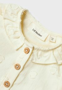 Blusa di cotone color crema con colletto arricciato, texture a fiori e bottoni in legno. Le etichette indicano la taglia e il marchio.