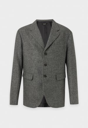 Homme portant un blazer gris, un T-shirt blanc imprimé, un pantalon noir, des chaussures noires, tenant un grand sac en cuir noir, debout devant un fond uni.