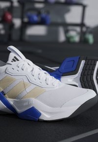 Scarpa da corsa Adidas bianca e beige con accenti blu poggiata su un pavimento nero della palestra con pesi sfocati sullo sfondo.