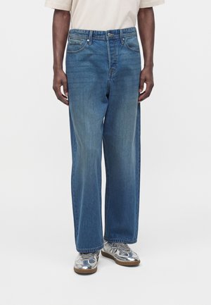 Personne portant un jean bleu à jambes larges, un t-shirt blanc et des baskets métalliques argentées, debout sur un fond clair uni.