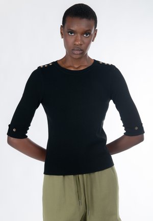 Vrouw draagt een zwart gebreid top met gouden knopen op de schouders en mouwen, gecombineerd met losse olijfgroene broek, staand tegen een witte achtergrond.