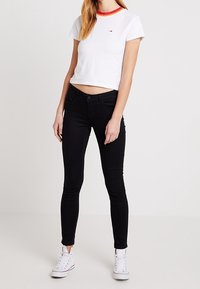 Mujer vestida con una camiseta corta blanca con cuello rojo y amarillo, jeans ajustados negros y zapatillas altas blancas, de pie contra un fondo blanco.