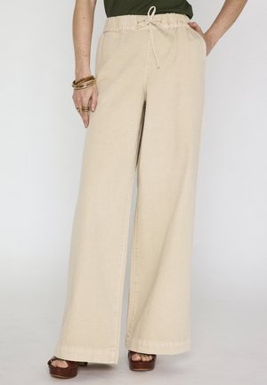 Pantalon beige à jambes larges avec ceinture élastique et cordon de serrage, porté avec des sandales compensées marron et des bracelets dorés au poignet gauche.