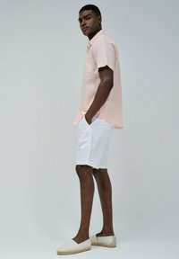 Camisa de manga corta rosa clara con un bolsillo en el pecho, combinada con pantalones cortos blancos y espadrilles a rayas beige en un modelo frente a un fondo neutro.