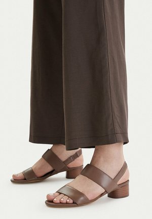 Piedi con sandali marroni con punta aperta, due cinturini e tacco basso a blocco, abbinati a pantaloni marroni a gamba larga.