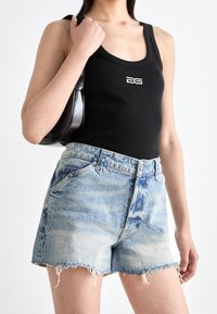 Kvinna i svart linne med vit logotyp, ljusblå fransade jeansshorts och bärandes en liten svart axelväska.