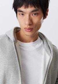 Grå zip-up hoodie med en blød tekstur og en kontrastfuld hvid t-shirt underneath. Fokus på hætten og den fulde lynlåsdetalje.