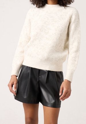 Femme portant un pull en maille texturée crème et un short plissé en cuir noir taille haute, debout avec les bras détendus le long du corps.