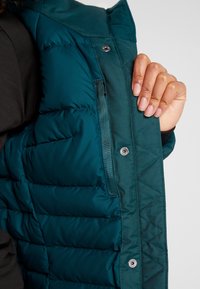 Mano sosteniendo abierta la parte delantera de una chaqueta acolchada color azul verdoso, mostrando botones de presión y un bolsillo interior.