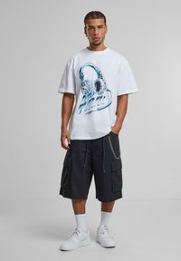 White short-sleeve t-shirt with blue headphone graphic, paired with black cargo shorts featuring pockets and chain detail, and white sneakers.