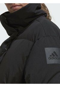Juodas pūkuotas švarkas su aukštu apykakle, turintis lygų paviršių. Su Adidas logotipo užsegimu ant rankovės ir mygtukų užsegimu.