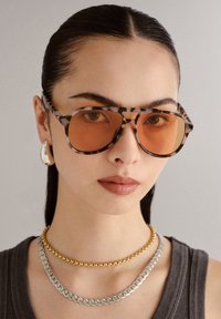 Brązowe okulary przeciwsłoneczne w kształcie skorupy żó�łwia z pomarańczowymi soczewkami, srebrne i złote naszyjniki łańcuchowe, szary top bez rękawów, gładka tekstura, oversize'owy geometryczny kształt.