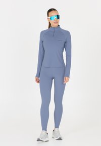 Helles blaues Sportoberteil mit halbem Reißverschluss, kombiniert mit passenden Leggings. Das Modell trägt reflektierende blaue Sonnenbrillen und graue Laufschuhe.