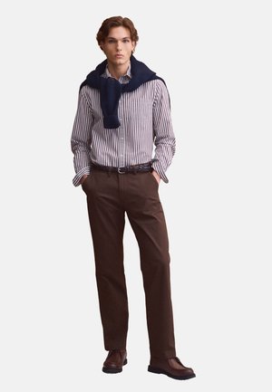 Giovane uomo che indossa una camicia a righe, pantaloni marroni, scarpe marroni e un maglione scuro drappeggiato sulle spalle, in piedi con le mani in tasca.